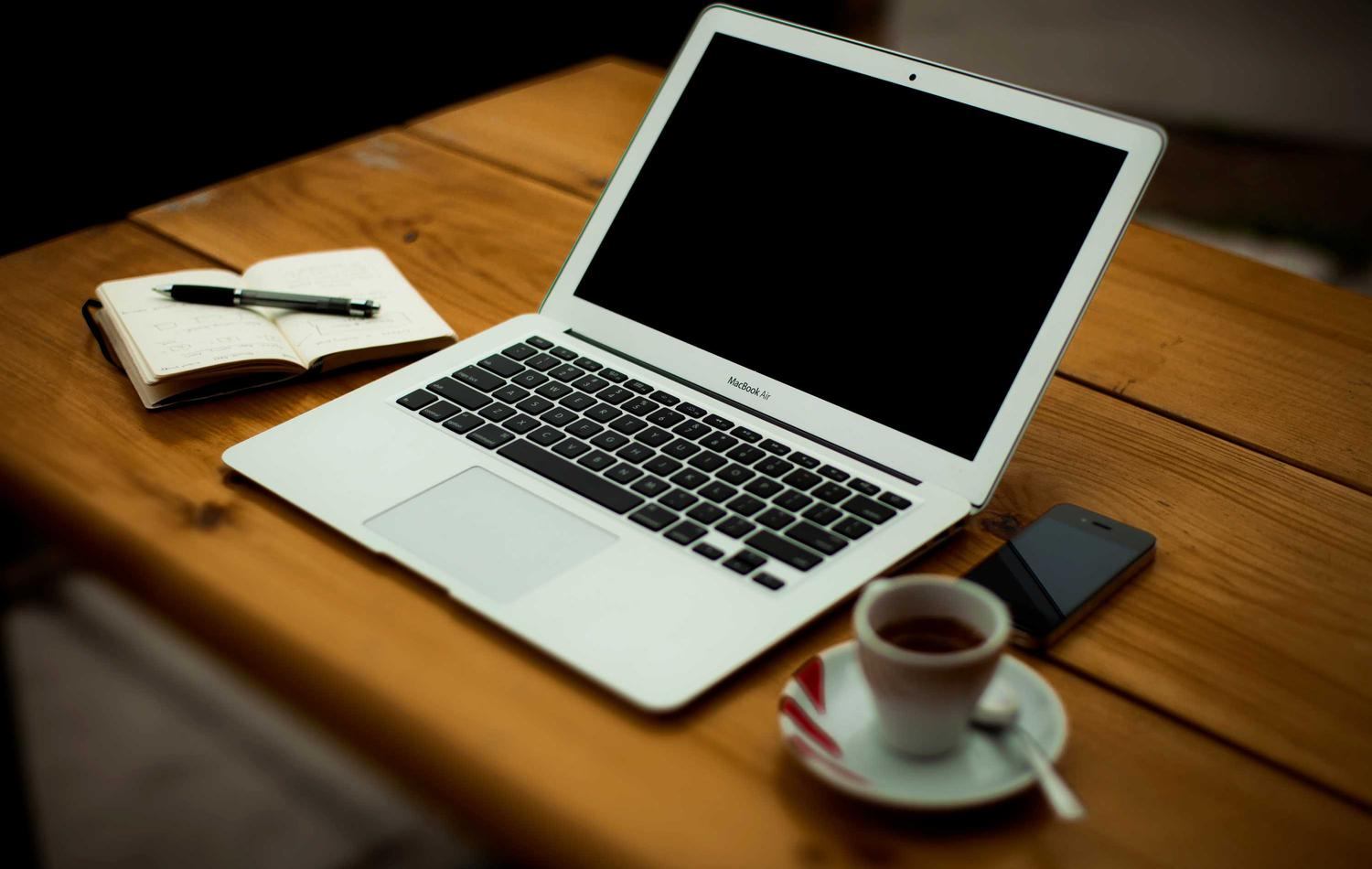 A laptop, notebook and coffee mug on a desk, ready for a working session.